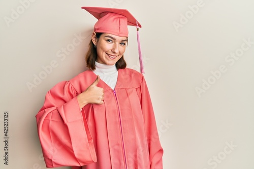 Young caucasian woman wearing graduation cap and ceremony robe doing happy thumbs up gesture with hand. approving expression looking at the camera showing success.
