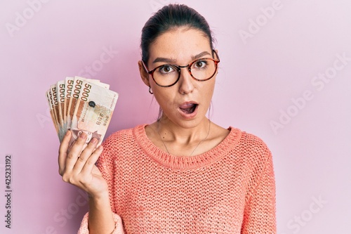 Young caucasian woman holding 5000 south korean won banknotes scared and amazed with open mouth for surprise, disbelief face