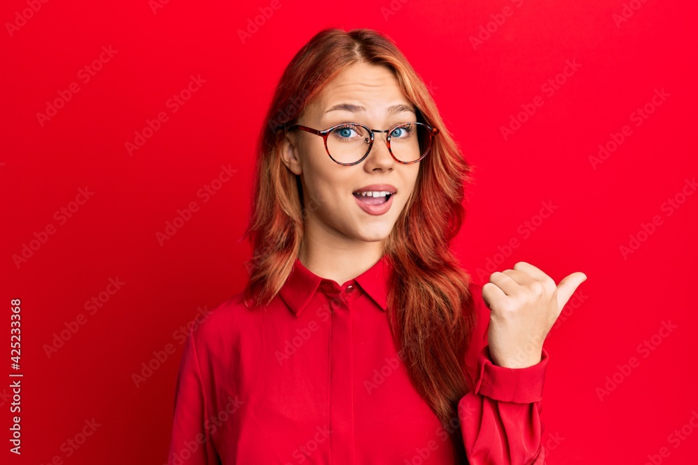 Young beautiful redhead woman wearing casual clothes and glasses over red background smiling with happy face looking and pointing to the side with thumb up. Young beautiful redhead woman wearing casual clothes and glasses over red background smiling with happy face looking and pointing to the side with thumb up.