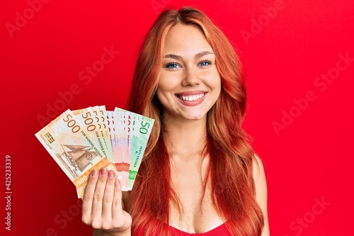Young beautiful redhead woman holding norwegian krone banknotes looking positive and happy standing and smiling with a confident smile showing teeth