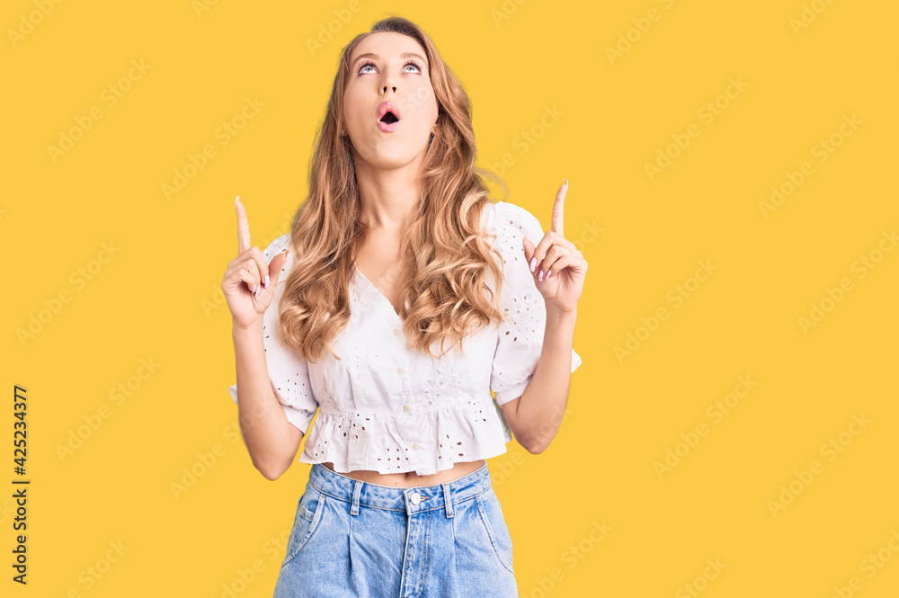 Young caucasian woman with blond hair wearing elegant summer tshirt amazed and surprised looking up and pointing with fingers and raised arms. Young caucasian woman with blond hair wearing elegant summer tshirt amazed and surprised looking up and pointing with fingers and raised arms.