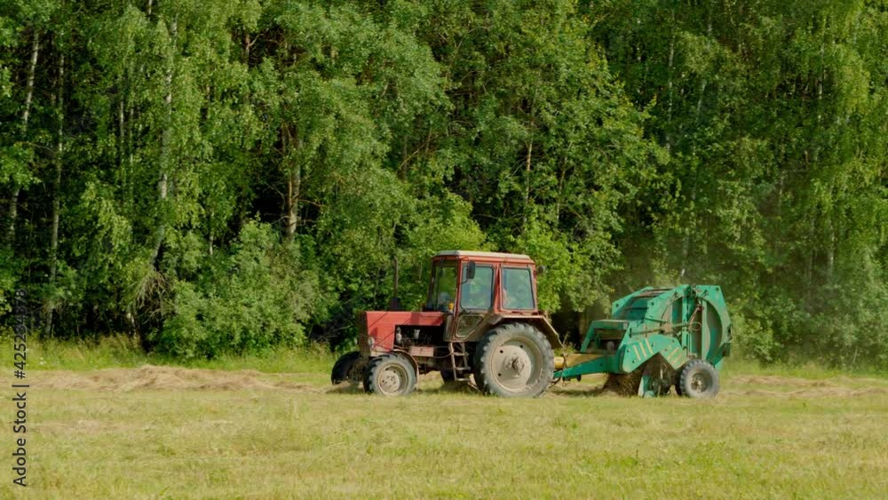 Harvesting hay. A farmer on a tractor with round baler rides on a wheat field after harvesting and makes hay bales. Agricultural machinery collects dry straw in rolls and round large bales. Harvesting hay. A farmer on a tractor with round baler rides on a wheat field after harvesting and makes hay bales. Agricultural machinery collects dry straw in rolls and round large bales.