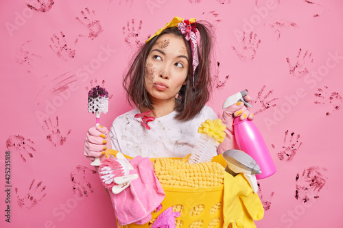 Serious thoughtful young Asian housewife holds detergent and brush looks pensively has dirty face poses near basket of laundry isolated over pink background with handprints. Housekeeping concept
