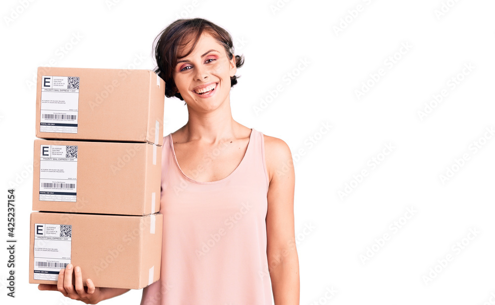 Beautiful young woman with short hair holding delivery packages looking positive and happy standing and smiling with a confident smile showing teeth Beautiful young woman with short hair holding delivery packages looking positive and happy standing and smiling with a confident smile showing teeth