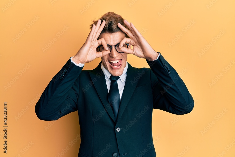 Handsome caucasian man wearing business suit and tie doing ok gesture like binoculars sticking tongue out, eyes looking through fingers. crazy expression. Handsome caucasian man wearing business suit and tie doing ok gesture like binoculars sticking tongue out, eyes looking through fingers. crazy expression.