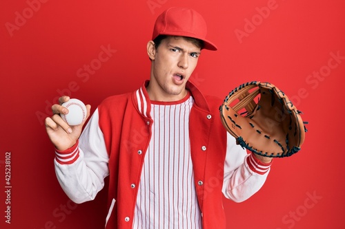 Handsome caucasian man wearing baseball uniform holding golve and ball in shock face, looking skeptical and sarcastic, surprised with open mouth photo