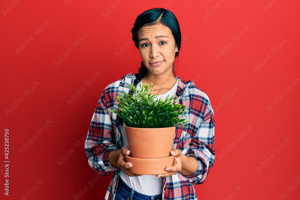 Beautiful hispanic woman wearing gardener shirt holding plant pot clueless and confused expression. doubt concept.