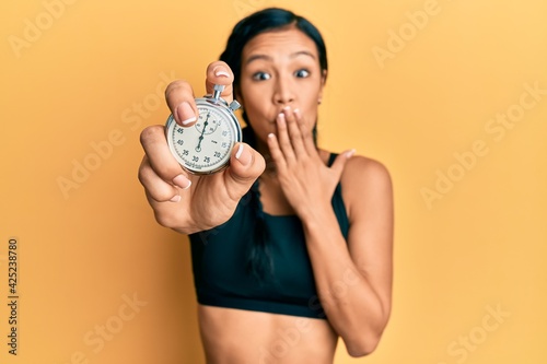 Beautiful hispanic woman wearing sportswear holding stopwatch covering mouth with hand  shocked and afraid for mistake. surprised expression