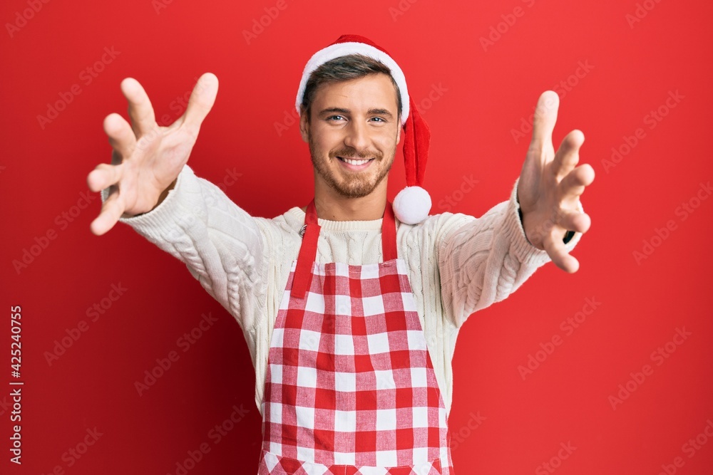 Handsome caucasian man wearing baker apron and christmas hat looking at the camera smiling with open arms for hug. cheerful expression embracing happiness. Handsome caucasian man wearing baker apron and christmas hat looking at the camera smiling with open arms for hug. cheerful expression embracing happiness.