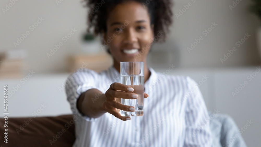Close up of happy young biracial woman stretch hand offer glass of clean still mineral water for body refreshment. Smiling African American female recommend aqua for hydration. Healthy life concept.