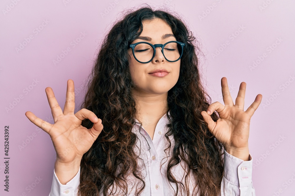 Young brunette woman with curly hair wearing casual clothes and glasses relax and smiling with eyes closed doing meditation gesture with fingers. yoga concept. Young brunette woman with curly hair wearing casual clothes and glasses relax and smiling with eyes closed doing meditation gesture with fingers. yoga concept.