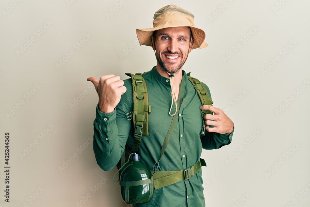 Handsome man with beard wearing explorer hat and backpack pointing to the back behind with hand and thumbs up, smiling confident Handsome man with beard wearing explorer hat and backpack pointing to the back behind with hand and thumbs up, smiling confident