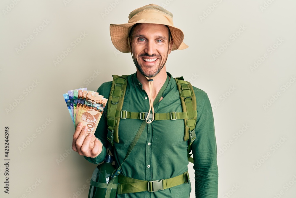 Handsome man with beard wearing explorer hat holding canadian dollars looking positive and happy standing and smiling with a confident smile showing teeth