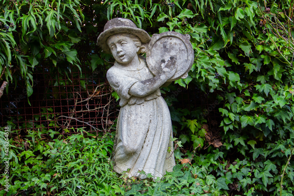 Old white weathered statue of a girl playing a tambourine surrounded with ivy Old white weathered statue of a girl playing a tambourine surrounded with ivy