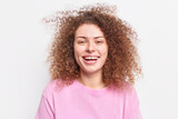 Horizontal shot of cheerful millennial girl smiles broadly shows teeth has curly bushy hair expresses joy being in good mood wears casual pink jumper isolated over white background. Emotions
