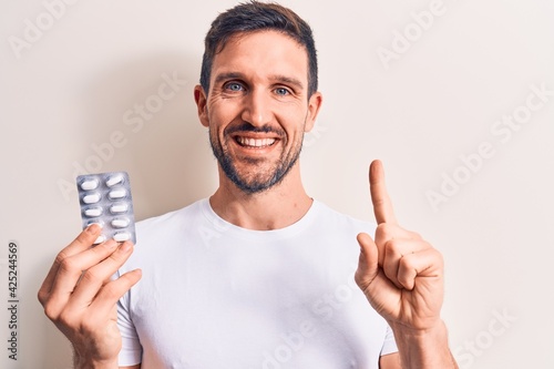 Young handsome man holding medicine pills standing over isolated white background smiling with an idea or question pointing finger with happy face, number one