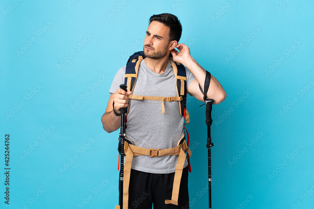 Young caucasian man with backpack and trekking poles isolated on blue background having doubts