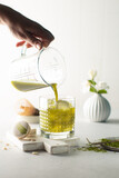 Women's hand pours green Matcha tea into the glass with ice in a white kitchen,with sweet macarons and ceramic vase on background.Close up of green tea.
