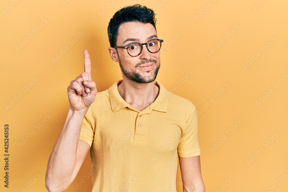 Fototapeta premium Young hispanic man wearing casual clothes and glasses showing and pointing up with finger number one while smiling confident and happy.