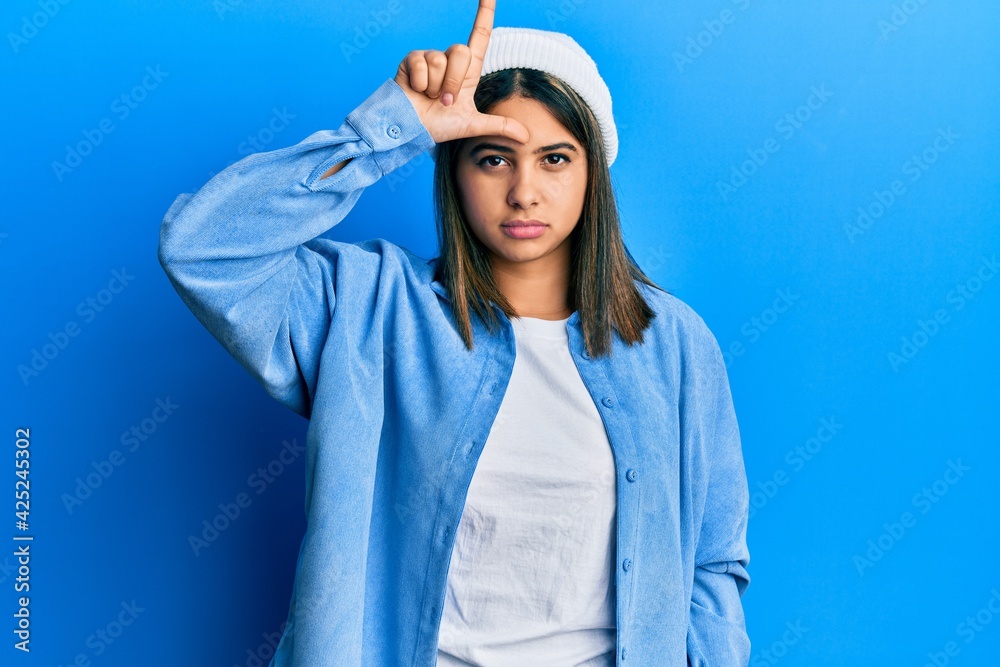 Young latin woman wearing cute wool cap making fun of people with fingers on forehead doing loser gesture mocking and insulting. Young latin woman wearing cute wool cap making fun of people with fingers on forehead doing loser gesture mocking and insulting.