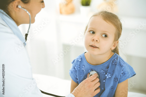 Doctor examining a child by stethoscope. Happy smiling girl patient dressed in blue dress is at usual medical inspection. Medicine concept