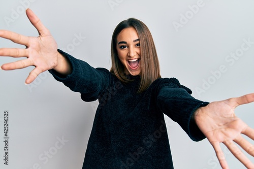 Young brunette girl wearing elegant fashion sweater looking at the camera smiling with open arms for hug. cheerful expression embracing happiness.