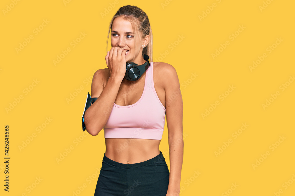 Young blonde woman wearing gym clothes and using headphones looking stressed and nervous with hands on mouth biting nails. anxiety problem. Young blonde woman wearing gym clothes and using headphones looking stressed and nervous with hands on mouth biting nails. anxiety problem.