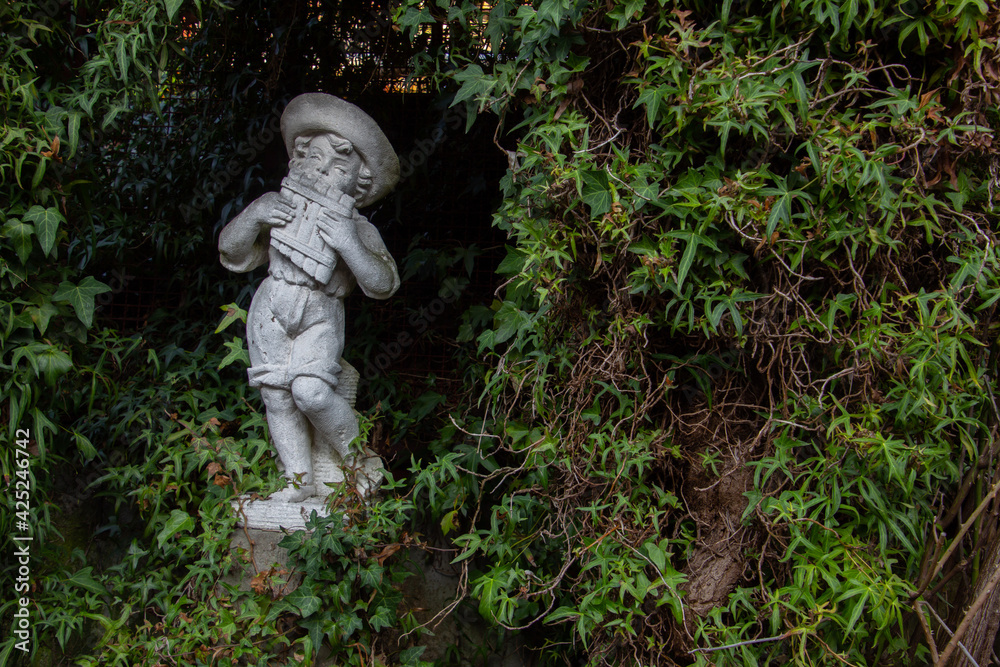 Old white weathered statue of a boy playing a pan flute surrounded with ivy