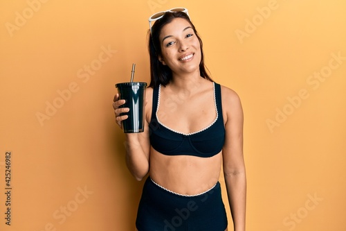 Young latin woman wearing bikini drinking soda looking positive and happy standing and smiling with a confident smile showing teeth © Krakenimages.com