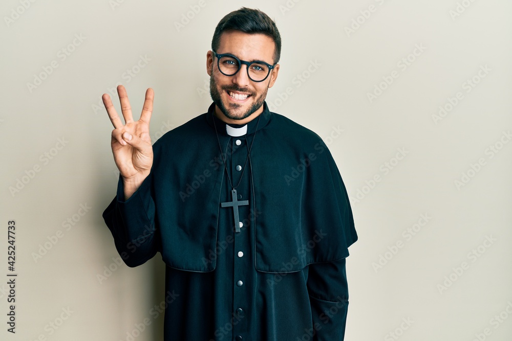 Young hispanic man wearing priest uniform standing over white background showing and pointing up with fingers number three while smiling confident and happy.
