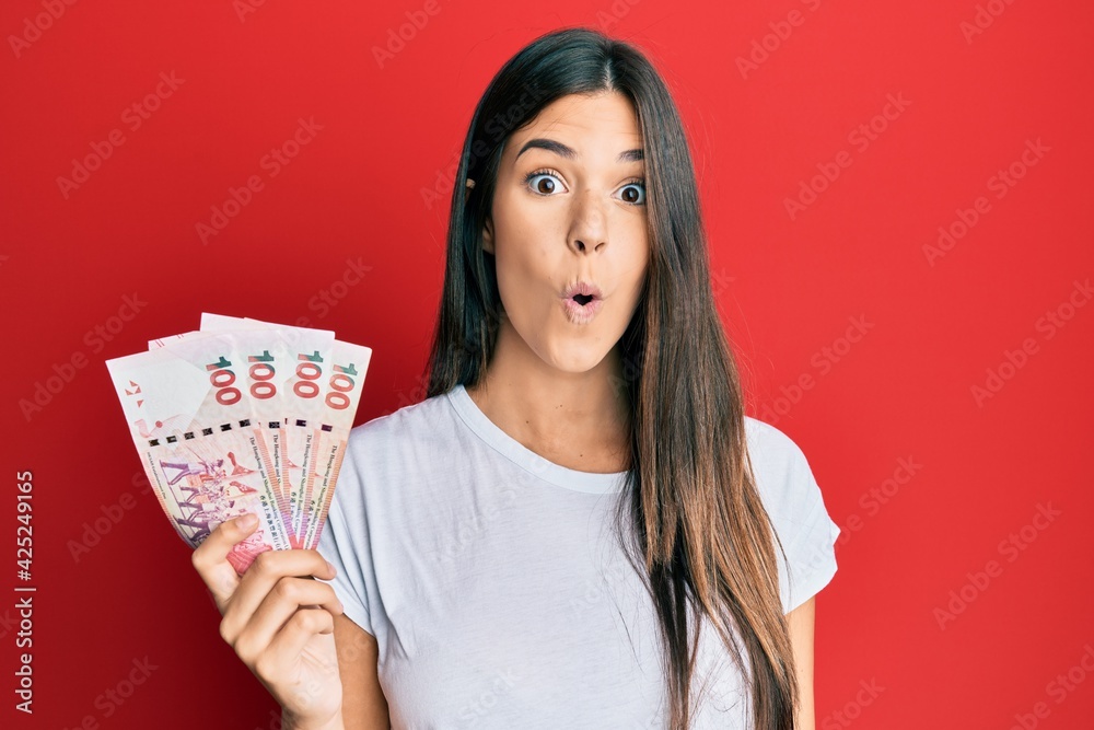 Young brunette woman holding hong kong 100 dollars banknotes scared and amazed with open mouth for surprise, disbelief face Young brunette woman holding hong kong 100 dollars banknotes scared and amazed with open mouth for surprise, disbelief face