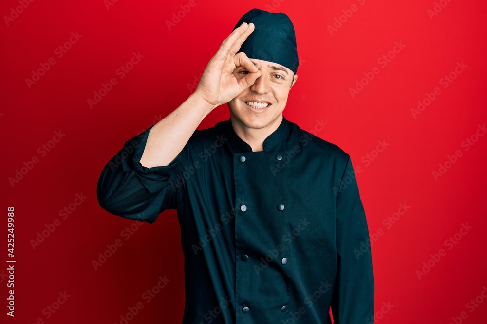 Handsome young man wearing professional cook uniform and hat doing ok gesture with hand smiling, eye looking through fingers with happy face. Handsome young man wearing professional cook uniform and hat doing ok gesture with hand smiling, eye looking through fingers with happy face.