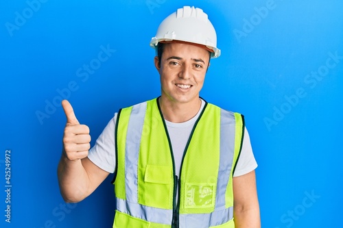 Handsome young man wearing safety helmet and reflective jacket smiling happy and positive, thumb up doing excellent and approval sign
