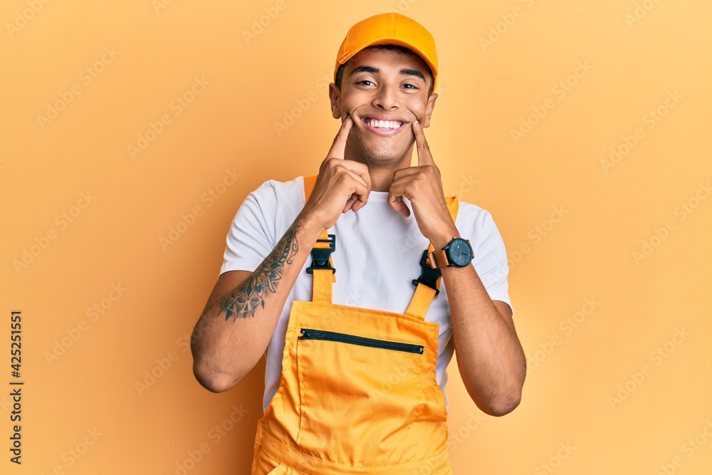 Young handsome african american man wearing handyman uniform over yellow background smiling with open mouth, fingers pointing and forcing cheerful smile
