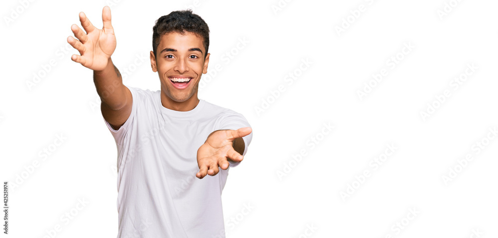 Young handsome african american man wearing casual white tshirt looking at the camera smiling with open arms for hug. cheerful expression embracing happiness. Young handsome african american man wearing casual white tshirt looking at the camera smiling with open arms for hug. cheerful expression embracing happiness.