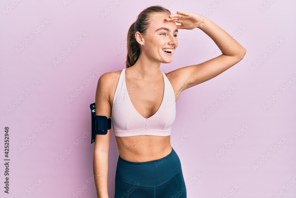 Beautiful blonde woman wearing sportswear and arm band very happy and smiling looking far away with hand over head. searching concept.
