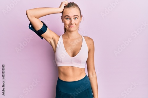 Beautiful blonde woman wearing sportswear and arm band confuse and wonder about question. uncertain with doubt, thinking with hand on head. pensive concept. © Krakenimages.com