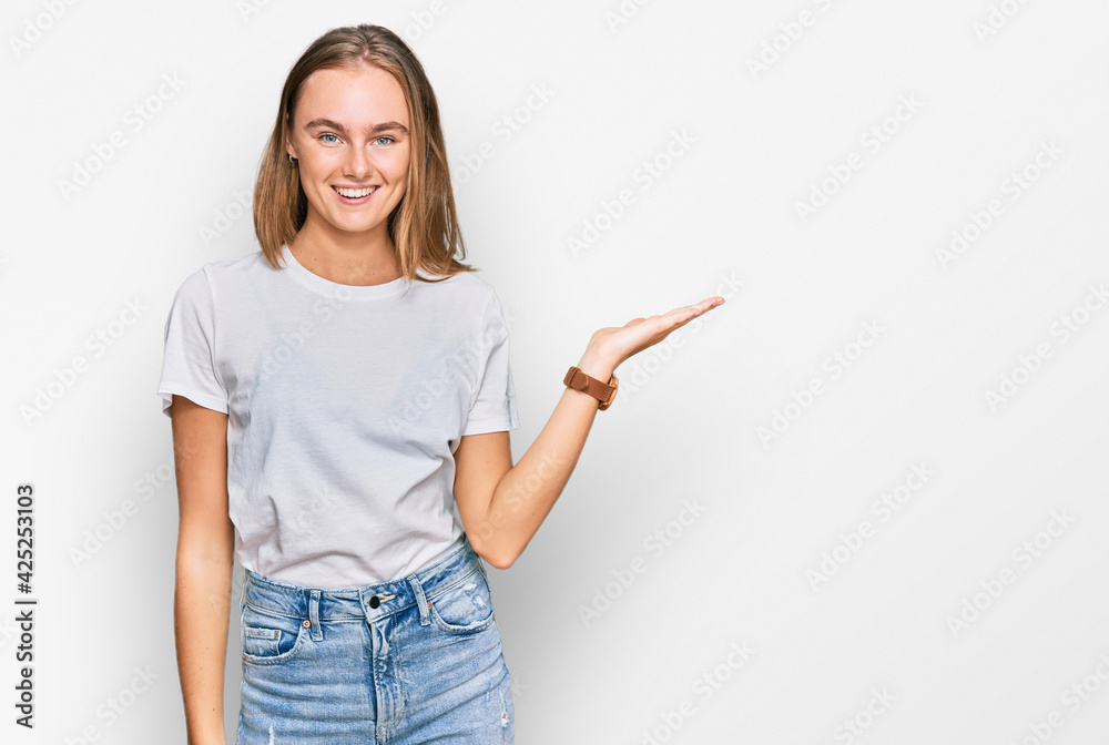 Beautiful young blonde woman wearing casual white t shirt smiling cheerful presenting and pointing with palm of hand looking at the camera. Beautiful young blonde woman wearing casual white t shirt smiling cheerful presenting and pointing with palm of hand looking at the camera.
