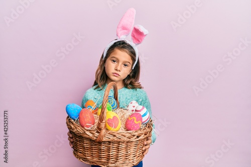Little beautiful girl wearing cute easter bunny ears holding wicker basket with colored eggs relaxed with serious expression on face. simple and natural looking at the camera.