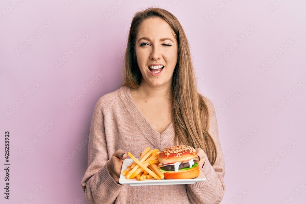 Young blonde woman eating a tasty classic burger with fries winking looking at the camera with sexy expression, cheerful and happy face. Young blonde woman eating a tasty classic burger with fries winking looking at the camera with sexy expression, cheerful and happy face.