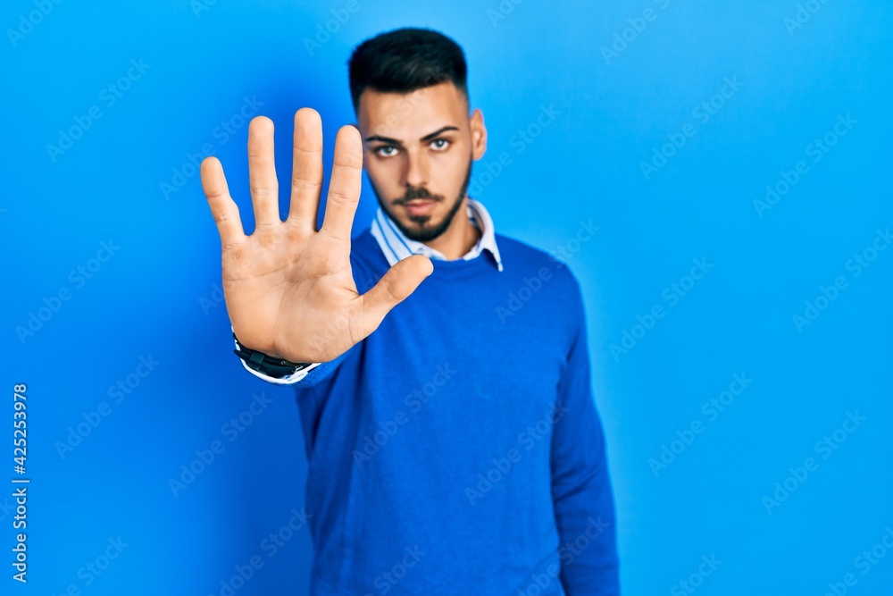 Fototapeta premium Young hispanic man with beard wearing casual blue sweater doing stop sing with palm of the hand. warning expression with negative and serious gesture on the face.