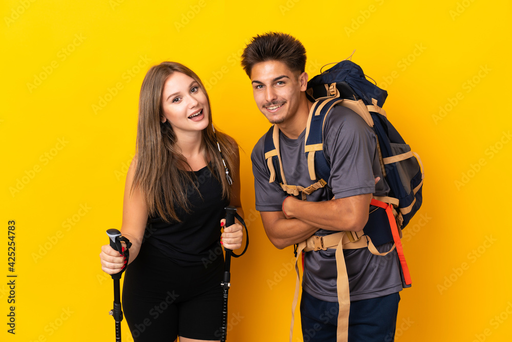 Young mountaineer couple with a big backpack isolated on yellow background keeping the arms crossed while smiling