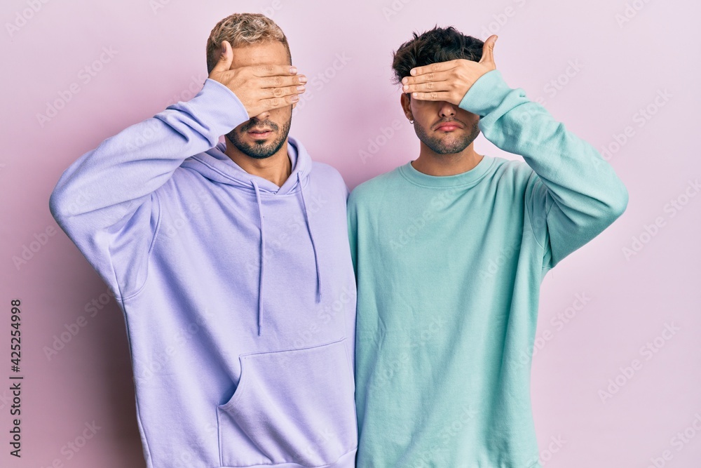 Homosexual gay couple standing together wearing casual clothes covering eyes with hand, looking serious and sad. sightless, hiding and rejection concept
