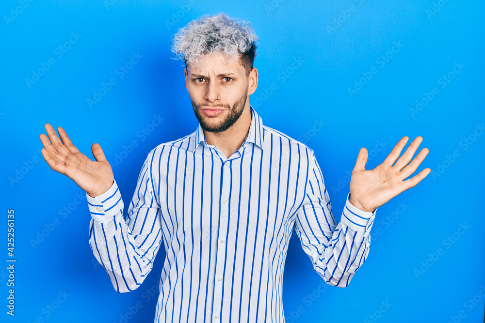 Naklejka premium Young hispanic man with modern dyed hair wearing business striped shirt clueless and confused with open arms, no idea and doubtful face.