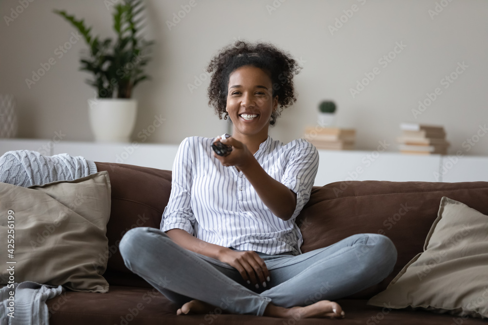 Overjoyed millennial African American female renter relax on couch in  living room watch TV online. Happy young biracial woman rest on sofa at  home have fun enjoying television program. Hobby concept. Stock