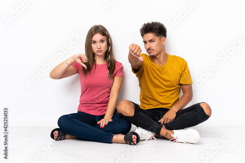 Young couple sitting on the floor isolated on white background showing thumb down with negative expression