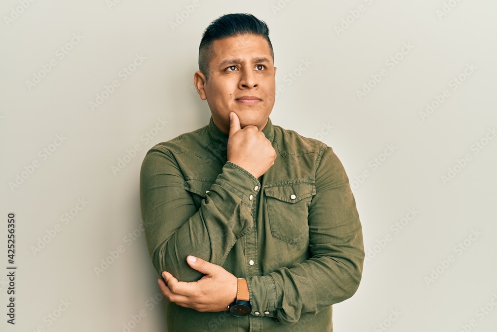 Young latin man wearing casual clothes with hand on chin thinking about question, pensive expression. smiling and thoughtful face. doubt concept. Young latin man wearing casual clothes with hand on chin thinking about question, pensive expression. smiling and thoughtful face. doubt concept.