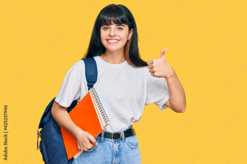 Young hispanic girl wearing student backpack and holding book smiling happy and positive, thumb up doing excellent and approval sign