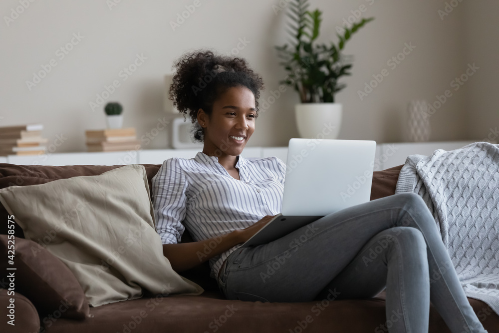 Happy millennial biracial female renter tenant rest on sofa in living room have fun using modern laptop gadget. Smiling young African American woman relax on couch at home browsing on computer. Happy millennial biracial female renter tenant rest on sofa in living room have fun using modern laptop gadget. Smiling young African American woman relax on couch at home browsing on computer.