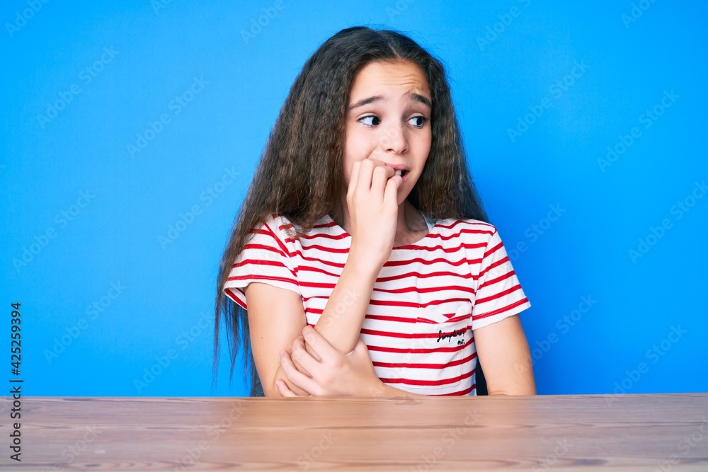 Cute hispanic child girl wearing casual clothes sitting on the table looking stressed and nervous with hands on mouth biting nails. anxiety problem. Cute hispanic child girl wearing casual clothes sitting on the table looking stressed and nervous with hands on mouth biting nails. anxiety problem.